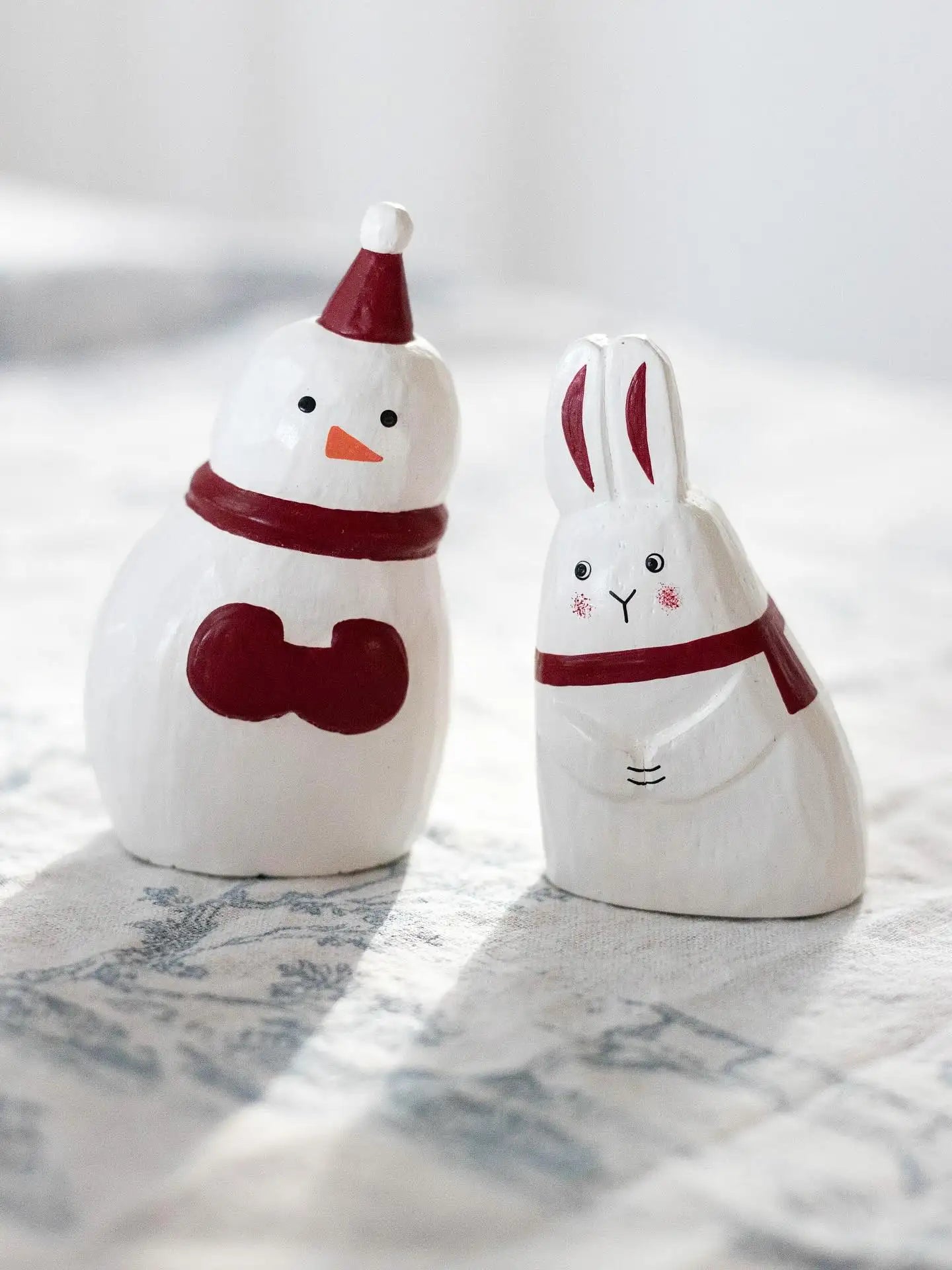 Figurines de bonhomme de neige et de lapin en céramique blanche, chacune ornée d'accents rouges incluant un bonnet de Père Noël pour le bonhomme de neige et des écharpes autour de leur cou.