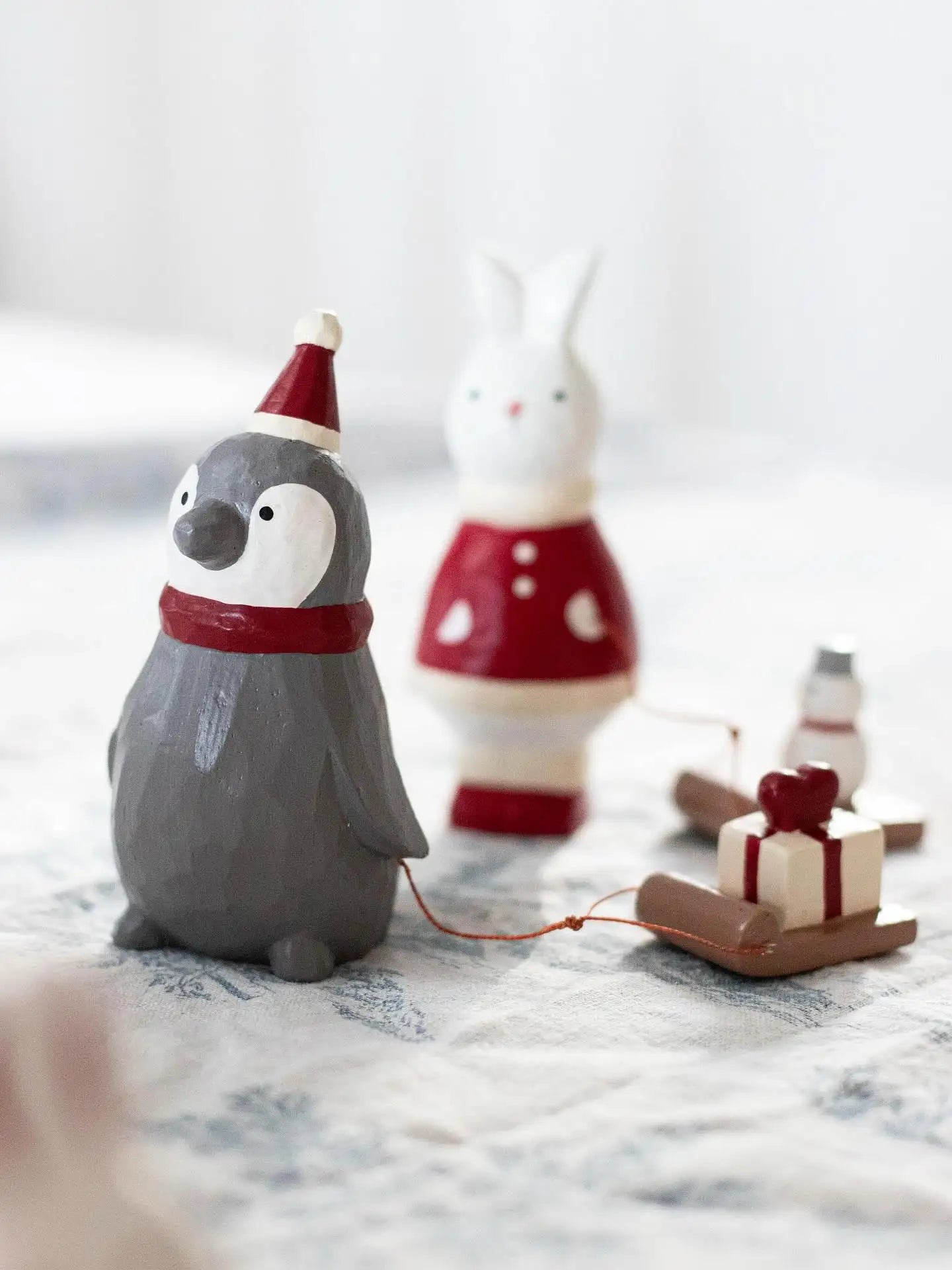 Petit pingouin en bois gris portant un bonnet de Père Noël rouge et une écharpe, tirant une petite luge avec une boîte cadeau.